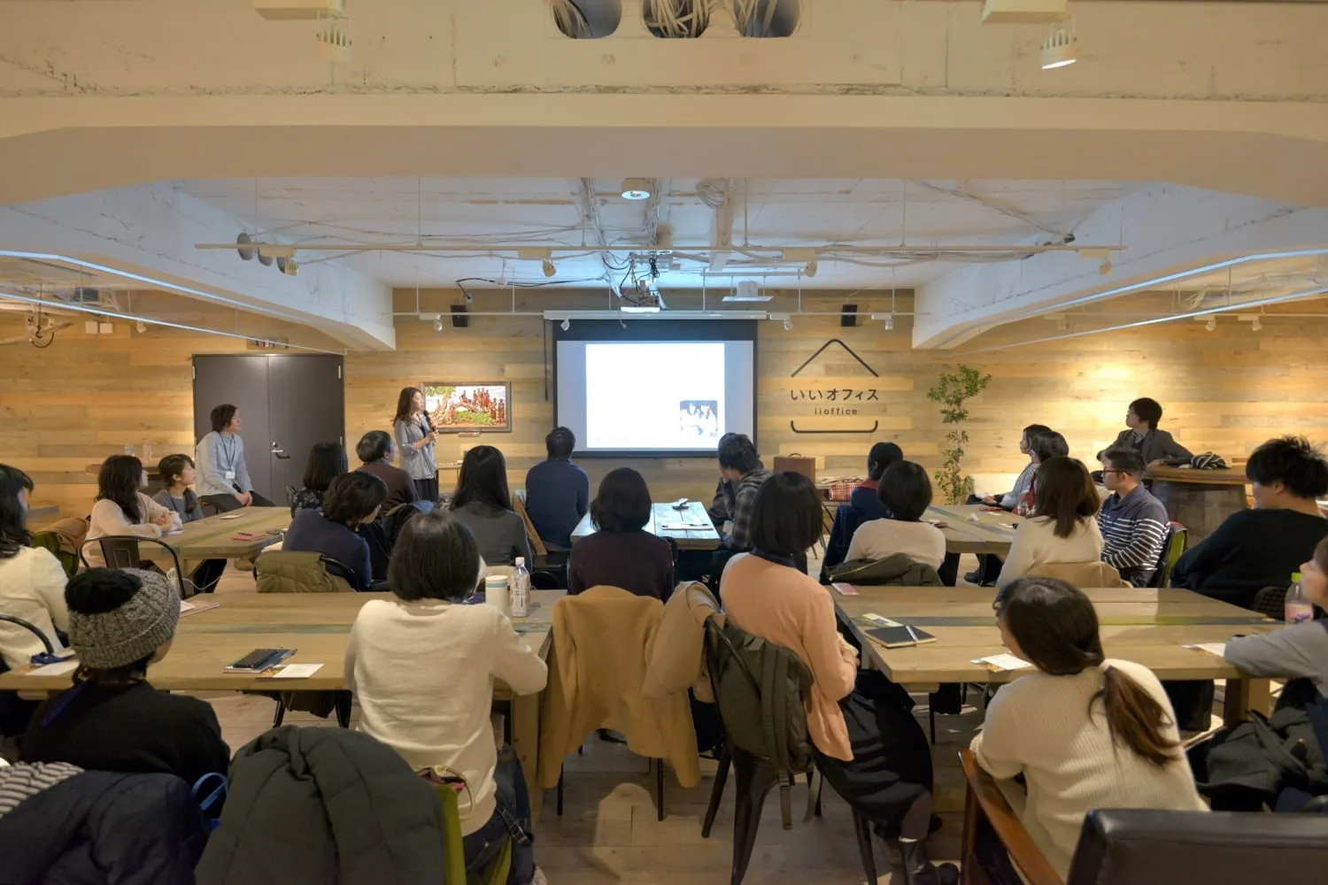 木の内装の会場で、テーブルに座って前の話を聞く参加者