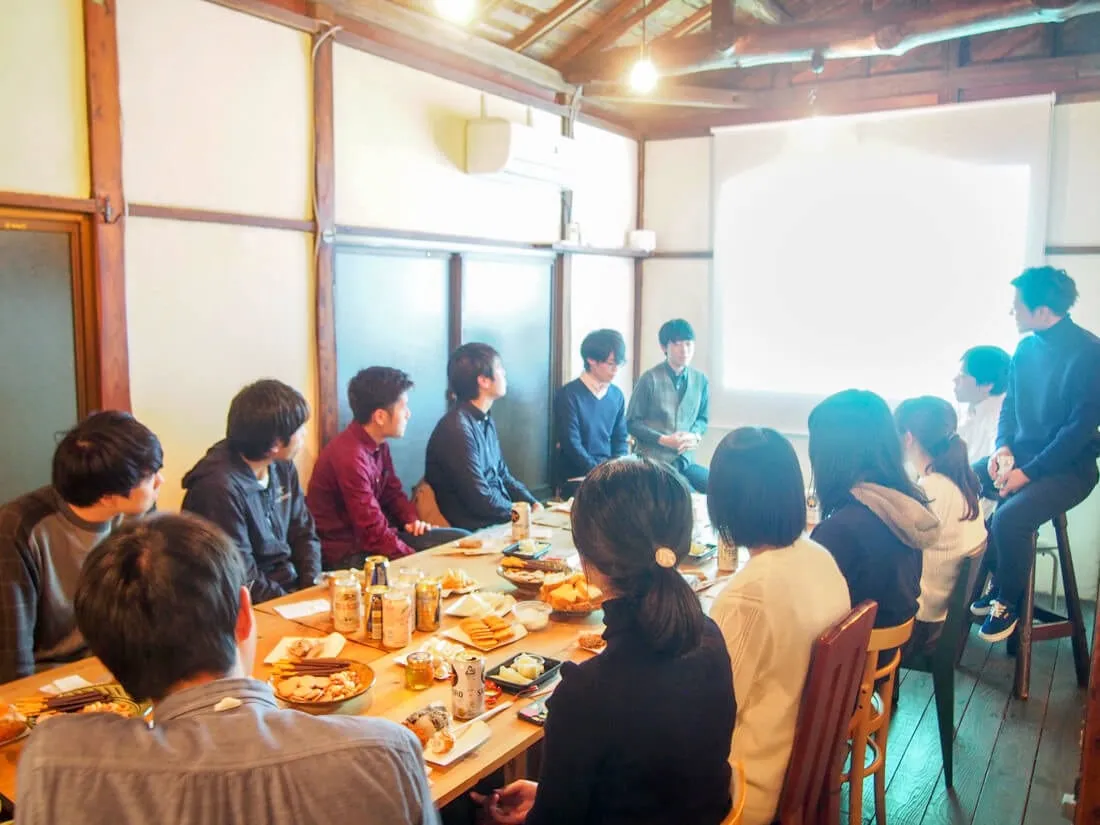 古い木造の部屋で長テーブルを囲み、食事をしながら話を聞く参加者